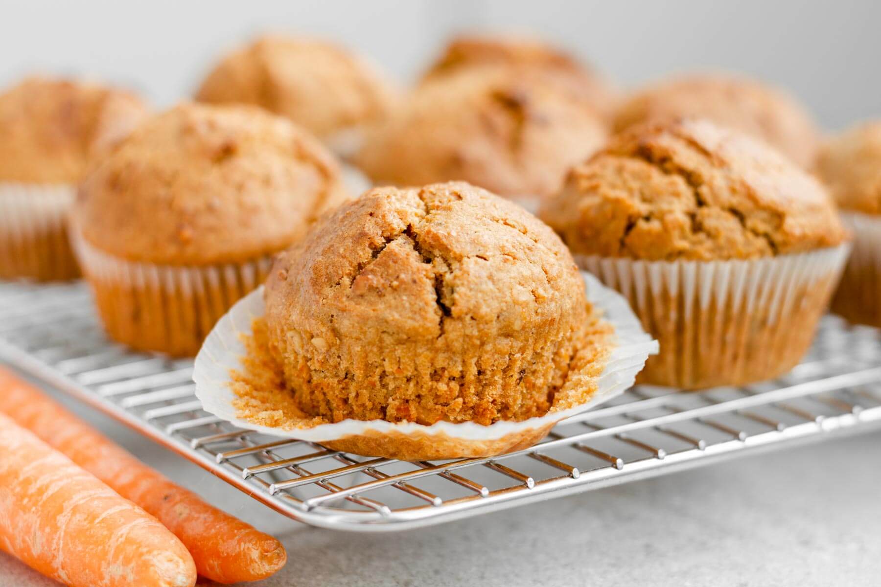 mehrere karottenmuffins auf kuchengitter und zwei karotten daneben.